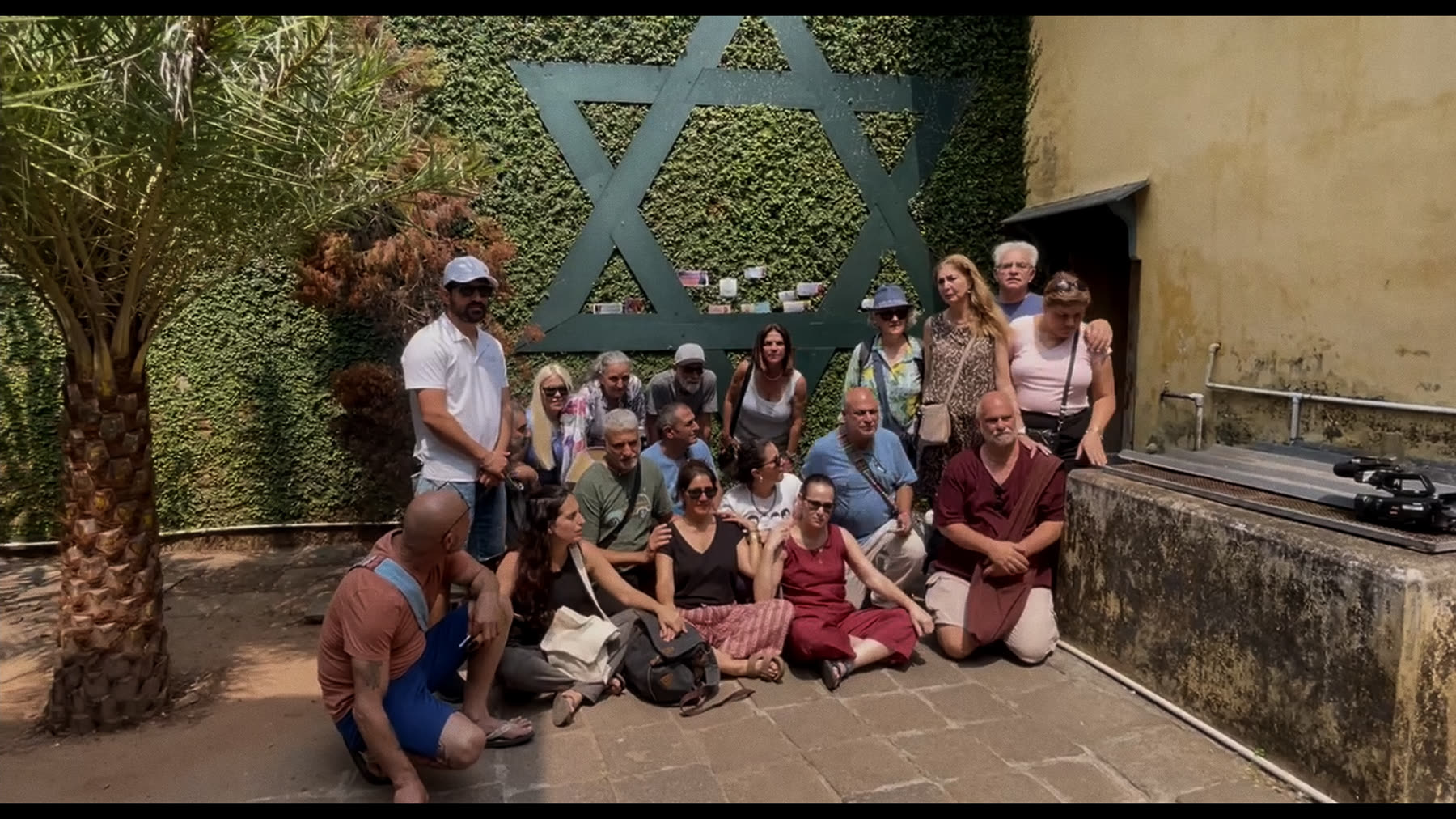 תצלום קבוצתי של הקבוצה בחצר המרחב, על רקע צמחייה וקיר אבן